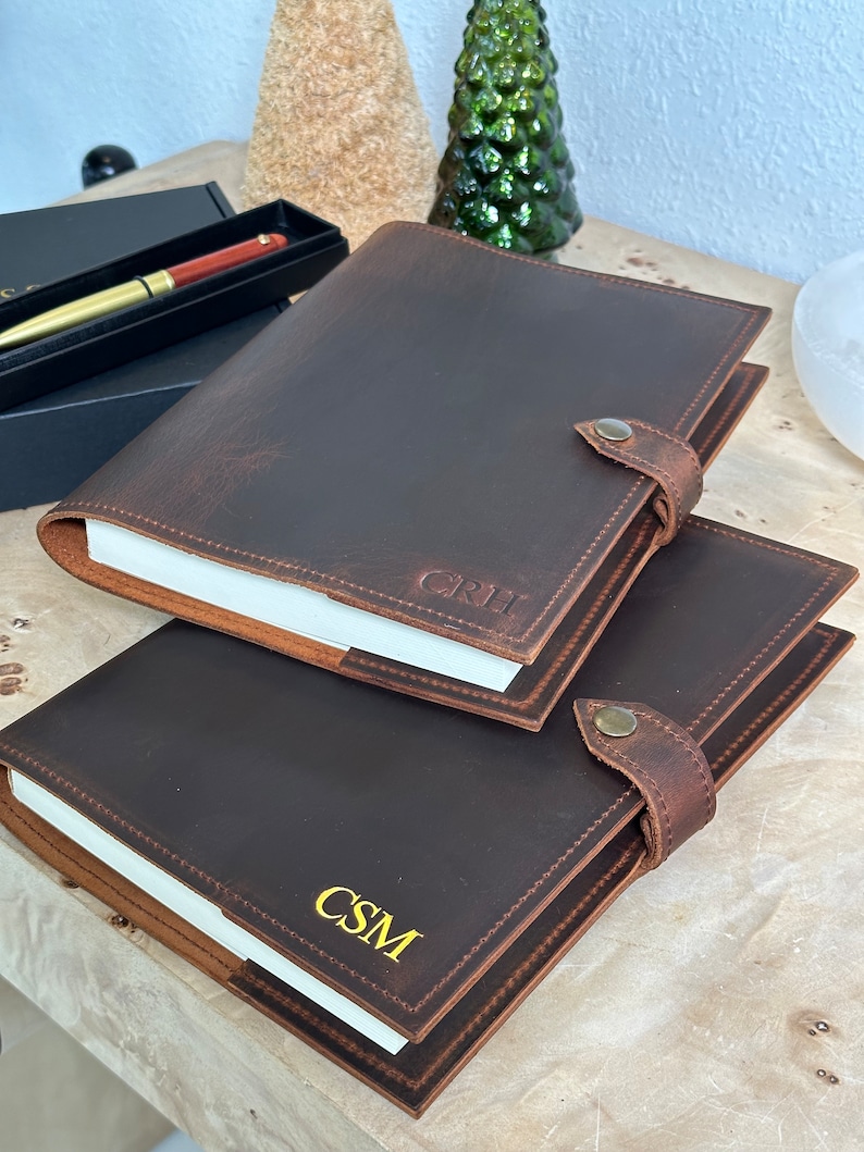 May include: Two brown leather journals with personalized initials, "CSM" and "GRH", in gold lettering. The journals have a flap closure with a metal button.