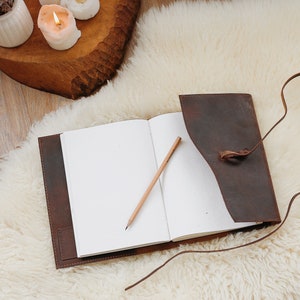 May include: A brown leather journal with a strap closure and a pencil lying open on a white fluffy surface.