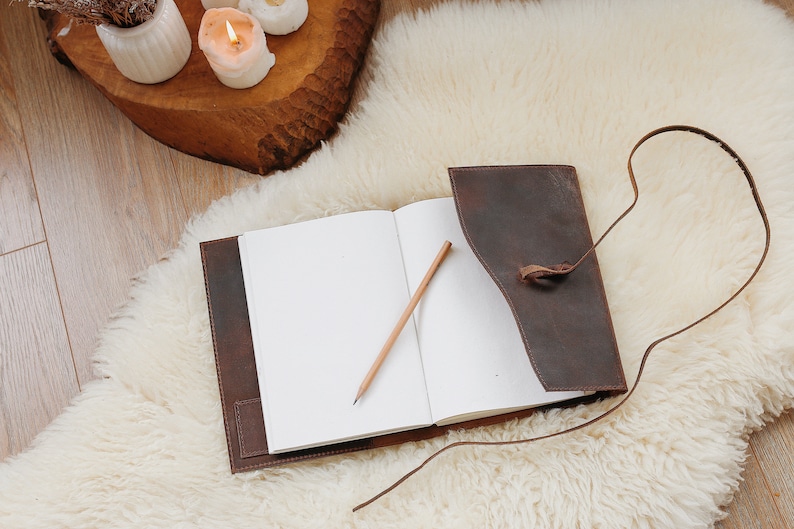 May include: A brown leather journal with a strap closure and a pencil lying open on a white fluffy surface.