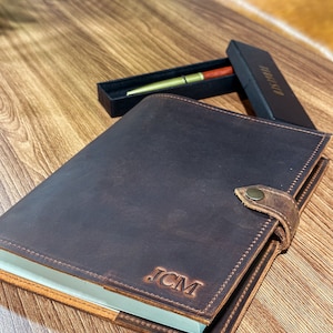 May include: A brown leather journal with a pen and a box. The journal has a flap closure and a strap with a button. The journal is personalized with the initials "JCM".