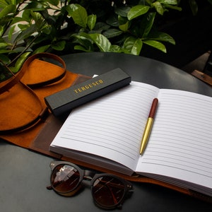 May include: A black leather journal with a gold pen and a black box with the text "TERGUSCO" on it. The journal is open to a blank page with lined paper. A pair of brown sunglasses is lying on the table.