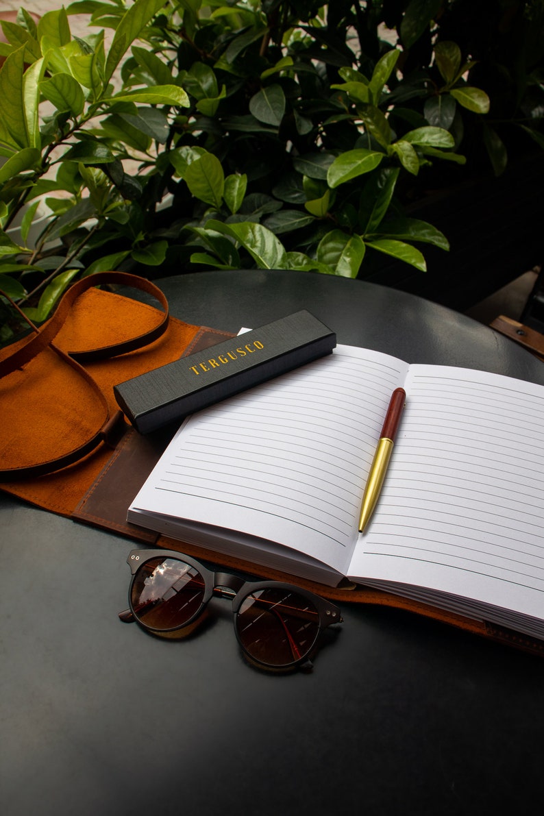 May include: A black leather journal with a gold pen and a black box with the text "TERGUSCO" on it. The journal is open to a blank page with lined paper. A pair of brown sunglasses is lying on the table.