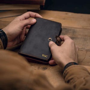 May include: A dark brown leather notebook cover featuring a snap button closure and the gold embossed initials TGC on the bottom right corner. The cover has visible stitching along the edges and a secure strap mechanism to keep the contents closed.