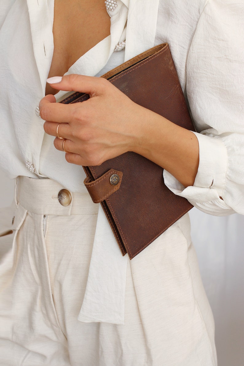 May include: A woman is holding a brown leather journal with a button closure. The journal is closed and has a smooth, textured surface.