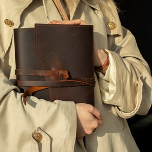 May include: A dark brown leather journal featuring a wrap-around leather cord closure. The journal has a smooth, matte finish with visible stitching along the edges and a knotted cord detail that secures the front cover in place.