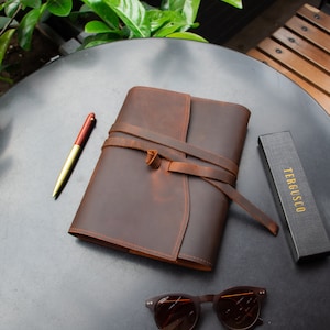 May include: A brown leather journal with a strap closure and a pen on a black table. The journal is tied with a brown leather strap. The journal is on a black table with a pair of brown sunglasses and a black box with the text "TERGUSCO" on it.