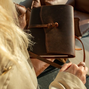 May include: A brown leather journal with a strap closure and a worn look. The journal is being held by a person wearing a beige jacket.