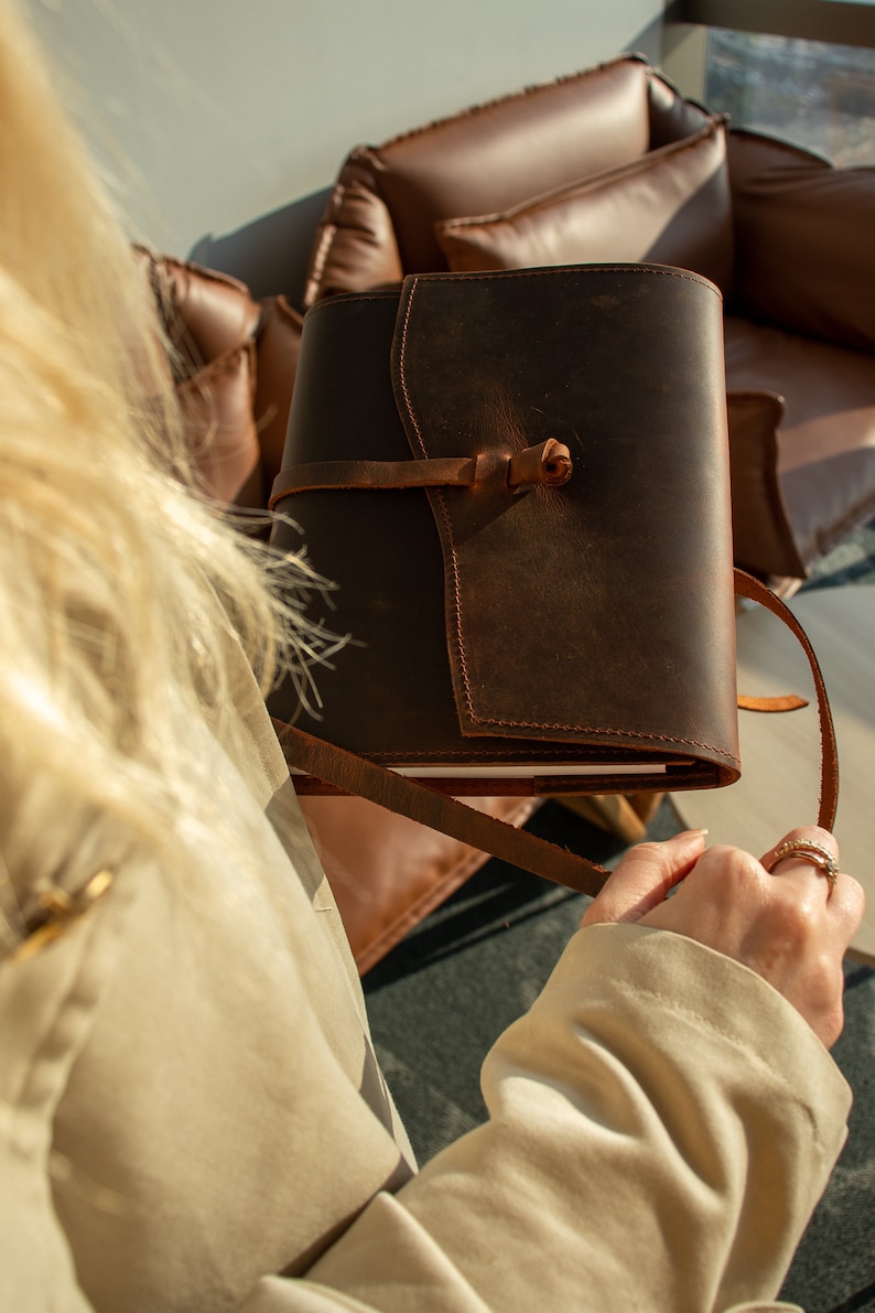 May include: A brown leather journal with a strap closure and a worn look. The journal is being held by a person wearing a beige jacket.