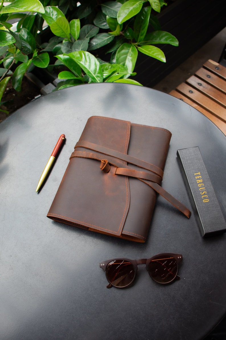 May include: A brown leather journal with a strap closure and a pen on a black table. The journal is tied with a brown leather strap. The journal is on a black table with a pair of brown sunglasses and a black box with the text "TERGUSCO" on it.