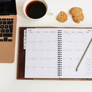 May include: A brown leather bound planner with a white monthly calendar for June 2022. A pencil is laying on the open planner. A laptop, a cup of coffee, and cookies are on a white surface.