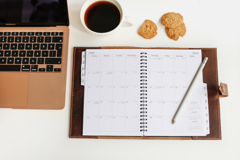 May include: A brown leather bound planner with a white monthly calendar for June 2022. A pencil is laying on the open planner. A laptop, a cup of coffee, and cookies are on a white surface.