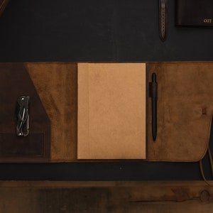 May include: A brown leather journal with a pen and a multi-tool in the pockets. The journal has a flap that closes with a strap.