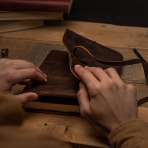 May include: A brown leather notebook cover with a flap that closes with a strap. The cover is open and the notebook pages are visible.