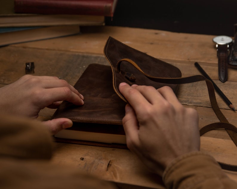 May include: A brown leather notebook cover with a flap that closes with a strap. The cover is open and the notebook pages are visible.