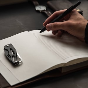 May include: A black pen is being used to write in a lined notebook with a brown leather cover. A small black multi-tool is lying on the open notebook.