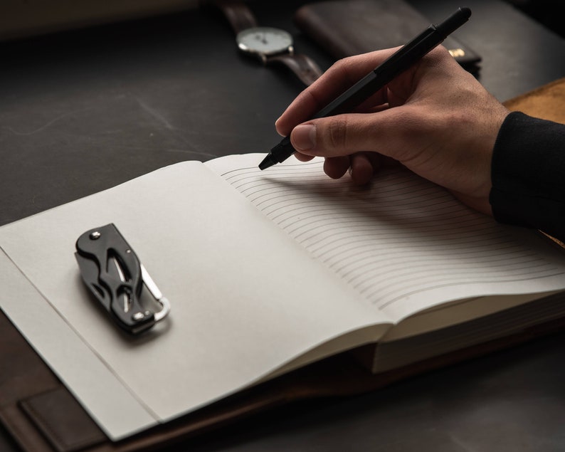 May include: A black pen is being used to write in a lined notebook with a brown leather cover. A small black multi-tool is lying on the open notebook.