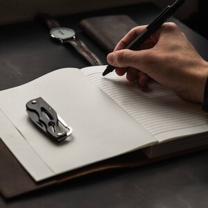 May include: A black pen is being used to write in a lined notebook. A small black pocket knife is on the left side of the notebook. A brown leather wallet and a wristwatch are in the background.