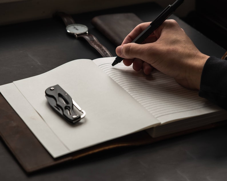 May include: A black pen is being used to write in a lined notebook. A small black pocket knife is on the left side of the notebook. A brown leather wallet and a wristwatch are in the background.