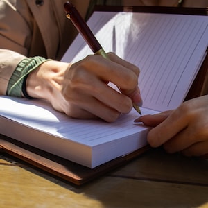 May include: A person writing in a brown leather bound journal with a gold and brown pen. The journal has lined white pages.