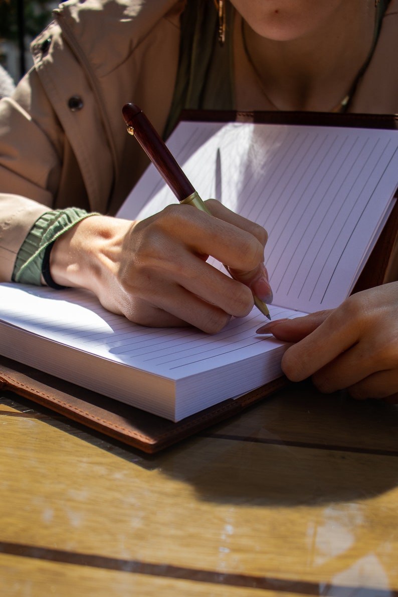 May include: A person writing in a brown leather bound journal with a gold and brown pen. The journal has lined white pages.