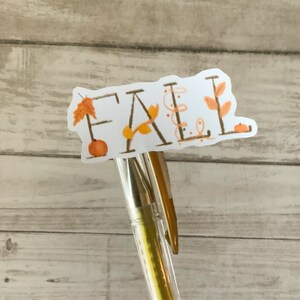 May include: A white sticker with the word "FALL" written in brown script. The sticker is decorated with small orange pumpkins, brown leaves, and brown vines. The sticker is attached to a gold pen.