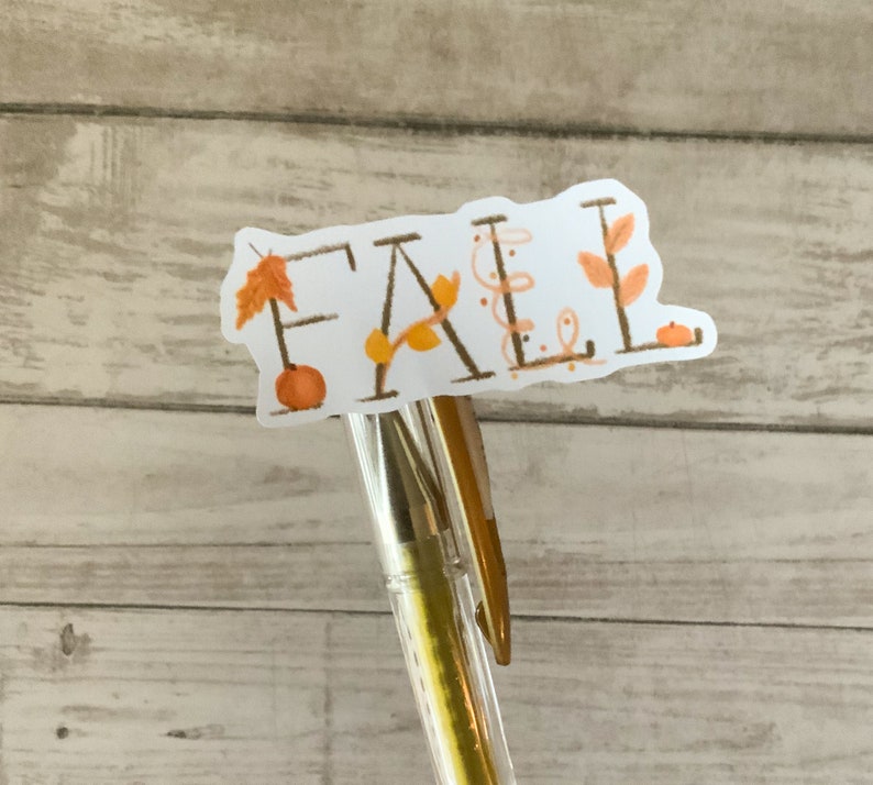 May include: A white sticker with the word "FALL" written in brown script. The sticker is decorated with small orange pumpkins, brown leaves, and brown vines. The sticker is attached to a gold pen.