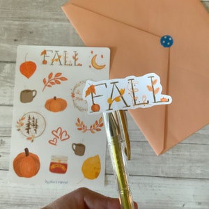 May include: A sheet of fall-themed stickers with illustrations of leaves, pumpkins, a forest, a cup of coffee, a jar of jam, and a leaf. The stickers are on a white background and have the text "FALL" in black lettering. The stickers are on a wooden surface with a peach-colored envelope in the background.