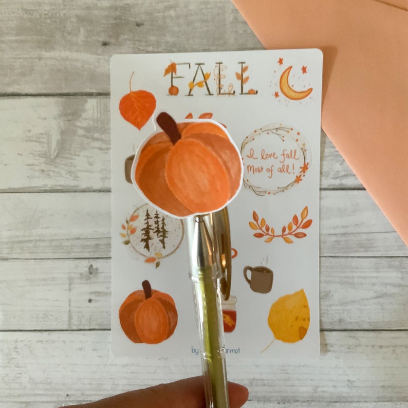 May include: A sheet of stickers featuring fall-themed illustrations, including pumpkins, leaves, a crescent moon, and a cup of coffee. The text "FALL" is written in large letters at the top of the sheet. The text "I love fall most of all!" is written in a circle with a wreath of branches.