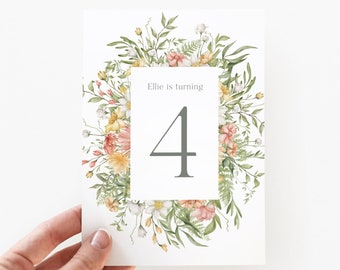 May include: A white card with a floral wreath border in shades of green, yellow, and pink. The text "Ellie is turning 4" is centered on the card.