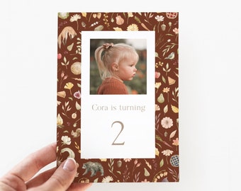 May include: A brown card with a watercolor pattern of fall leaves, berries, and other nature elements. A photo of a young girl is in the center of the card. The text "Cora is turning 2" is below the photo.