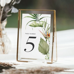 May include: A gold-framed table number card with a white background and tropical leaf and flower illustrations. The card reads "Table Number 5".