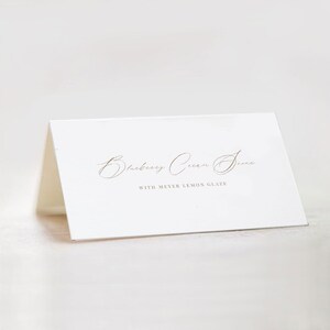 May include: White card with gold lettering that reads "Blueberry Cream Scone with Meyer Lemon Glaze".