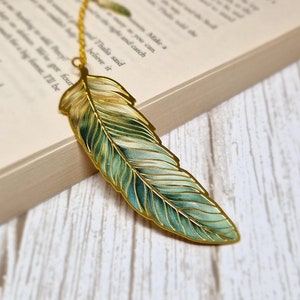 May include: A gold-coloured metal bookmark shaped like a feather with green and white enamel detailing.