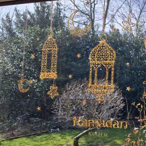 Op de afbeelding: Een raam versierd met gouden Ramadan-decoraties. De decoraties omvatten hangende lantaarns, halve manen en sterren. De woorden "Ramadan Kareem" staan in gouden letters onderaan het raam.