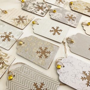 May include: A collection of 12 gift tags with a snowflake design. The tags are made of brown cardstock with a white background and have a gold glitter finish. Each tag has a small gold bell attached to the top.