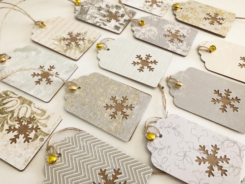 May include: A collection of 12 gift tags with a snowflake design. The tags are made of brown cardstock with a white background and have a gold glitter finish. Each tag has a small gold bell attached to the top.