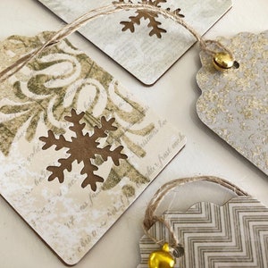 May include: Set of four gift tags with a rustic, vintage aesthetic. Each tag features a different design with snowflakes, a floral pattern, and a chevron pattern. The tags are made of brown cardstock and have a distressed look. Each tag has a gold bell attached to a twine string.