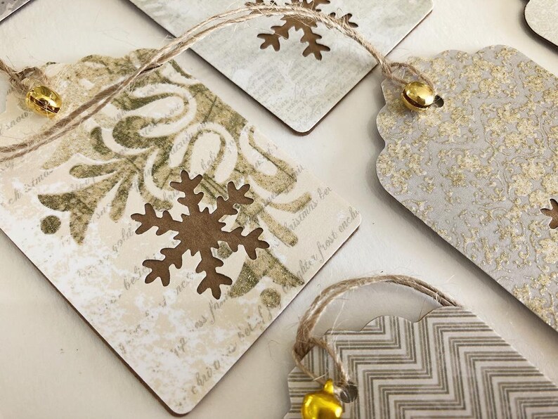 May include: Set of four gift tags with a rustic, vintage aesthetic. Each tag features a different design with snowflakes, a floral pattern, and a chevron pattern. The tags are made of brown cardstock and have a distressed look. Each tag has a gold bell attached to a twine string.
