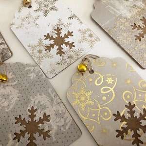 May include: Five gift tags with a snowflake design. The tags are made of brown paper with a white background and gold accents. Each tag has a small gold bell attached to it.
