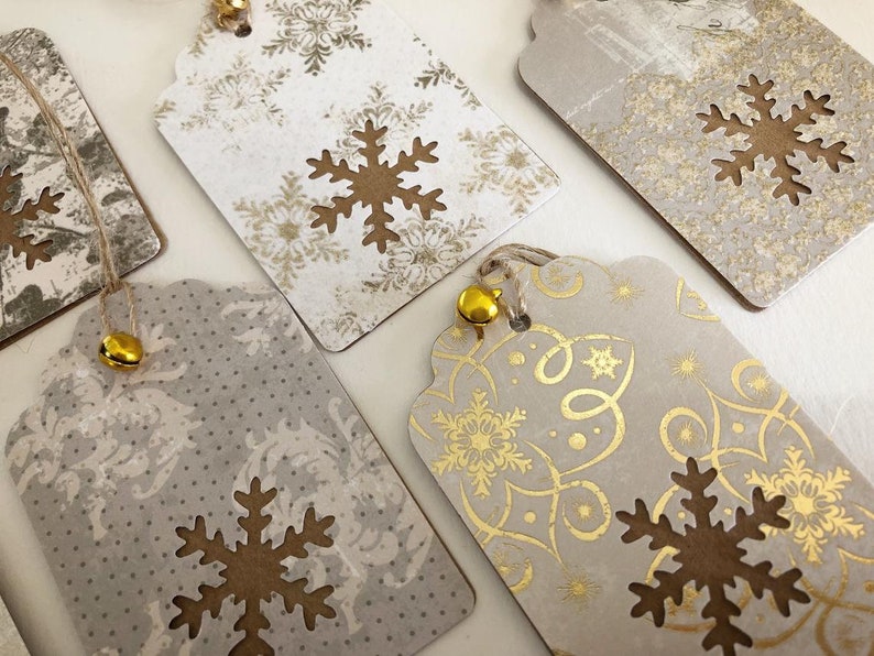 May include: Five gift tags with a snowflake design. The tags are made of brown paper with a white background and gold accents. Each tag has a small gold bell attached to it.