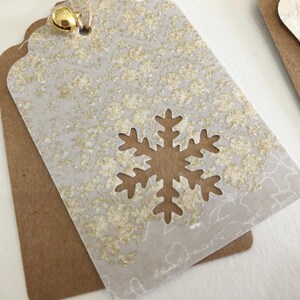 May include: A white gift tag with a gold snowflake cutout and a gold glitter pattern. The tag is attached to a brown paper gift tag with a gold bell.