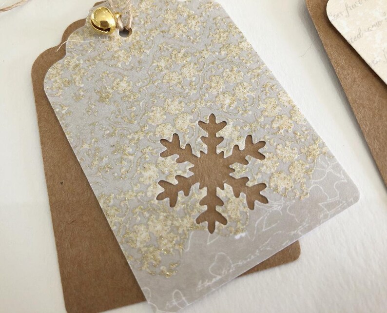 May include: A white gift tag with a gold snowflake cutout and a gold glitter pattern. The tag is attached to a brown paper gift tag with a gold bell.