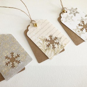 May include: Three white gift tags with gold snowflake designs and brown backing. Each tag has a gold bell attached to a twine string.