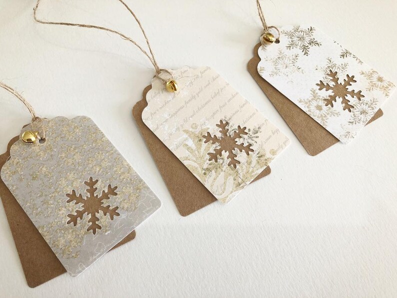 May include: Three white gift tags with gold snowflake designs and brown backing. Each tag has a gold bell attached to a twine string.
