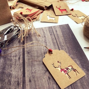 K&ouml;nnte beinhalten: Ein braunes Papier-Geschenk-Etikett mit einem ausgeschnittenen Rentier-Design. Das Rentier ist mit einem rot-gr&uuml;nen Karomuster verziert. Das Etikett ist mit einer Schnur gebunden und hat eine rote Perle.