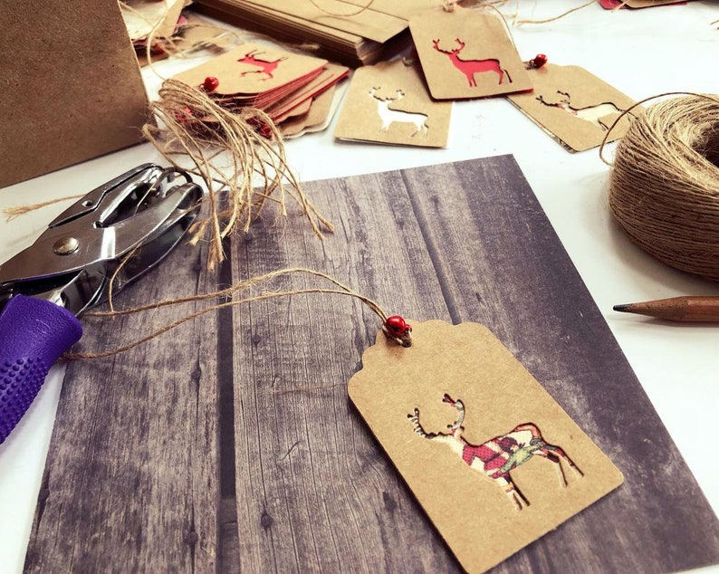 K&ouml;nnte beinhalten: Ein braunes Papier-Geschenk-Etikett mit einem ausgeschnittenen Rentier-Design. Das Rentier ist mit einem rot-gr&uuml;nen Karomuster verziert. Das Etikett ist mit einer Schnur gebunden und hat eine rote Perle.