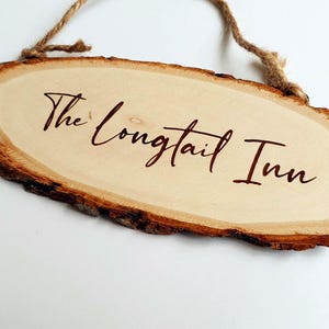 Può includere: Un cartello in legno con un bordo rustico in corteccia naturale, appeso a una corda di spago. Il cartello è di forma ovale e presenta il testo "The Longtail Inn" scritto in lettere corsive marroni.