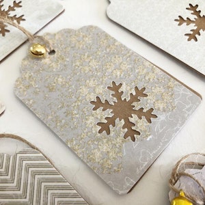 May include: A set of four gift tags with a snowflake design. The tags are made of white cardstock with a gold glitter finish. Each tag has a brown snowflake cut out of the center. The tags are tied with twine and have a small gold bell attached.