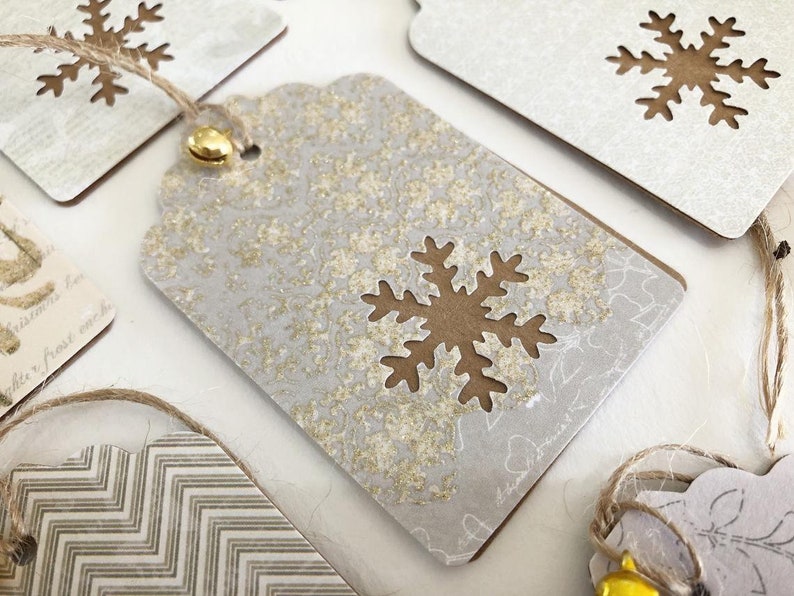 May include: A set of four gift tags with a snowflake design. The tags are made of white cardstock with a gold glitter finish. Each tag has a brown snowflake cut out of the center. The tags are tied with twine and have a small gold bell attached.