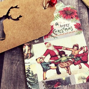 K&ouml;nnte beinhalten: Ein Weihnachtsgeschenkanh&auml;nger im Vintage-Stil mit braunem Papierhintergrund und einem ausgeschnittenen Rentiermotiv. Die Vorderseite des Anh&auml;ngers zeigt eine farbenfrohe Illustration von Weihnachtsmann und Kindern, die sich im Kreis an den H&auml;nden halten, mit dem Text "A happy Christmas" in einer dekorativen Schrift.